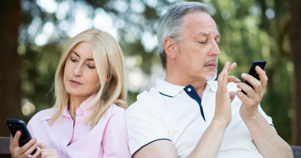 Older couple sitting back to back outdoors, each looking at their own phone