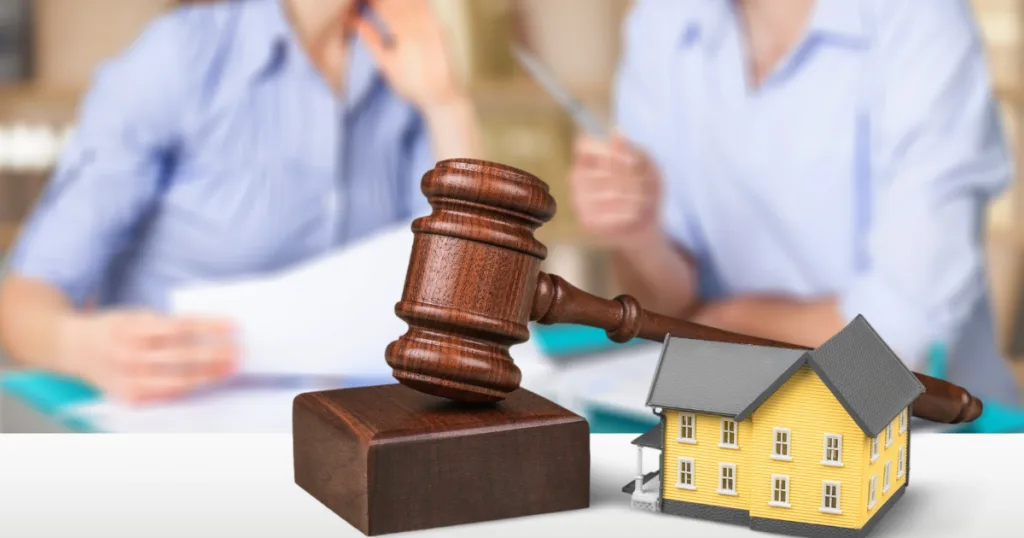 Wooden gavel next to a model house with a couple reviewing documents behind