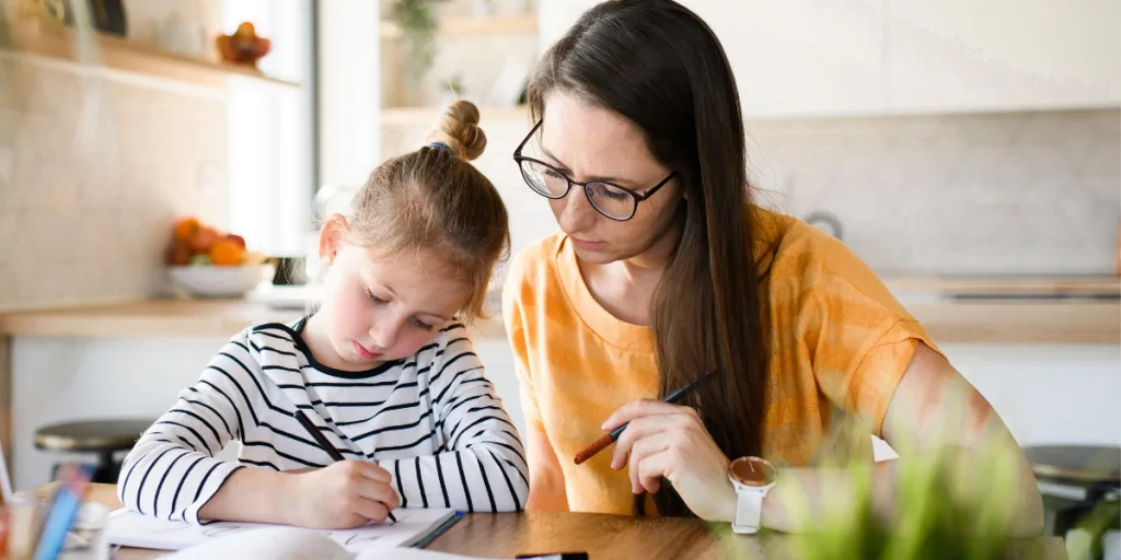 Mother in glasses helping her young daughter with homework at the kitchen table
