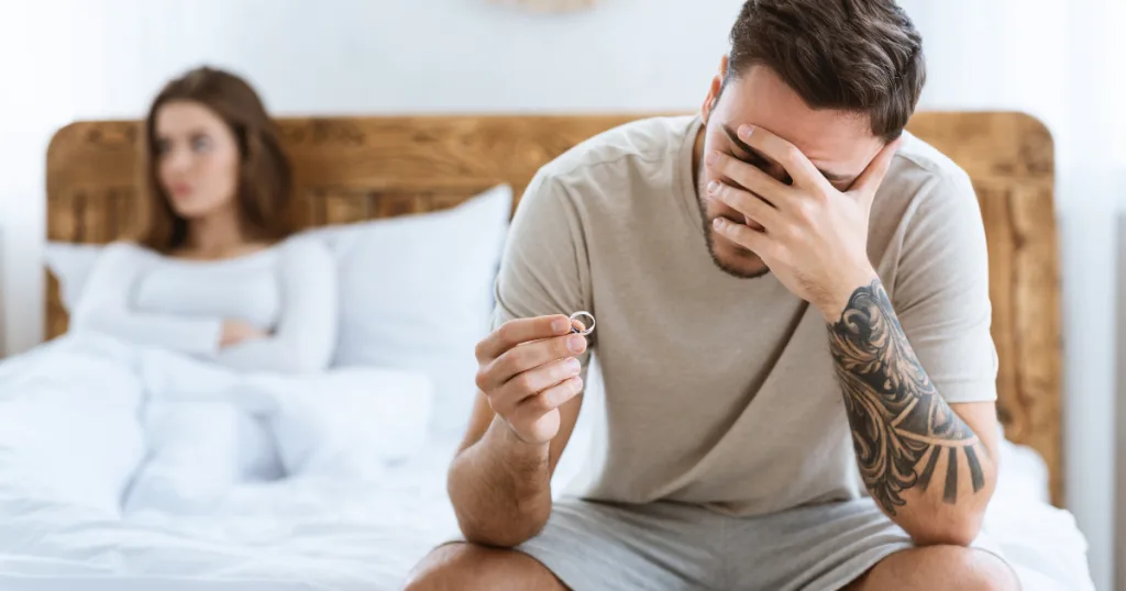 Distressed man holding a wedding ring on a bed with woman behind him
