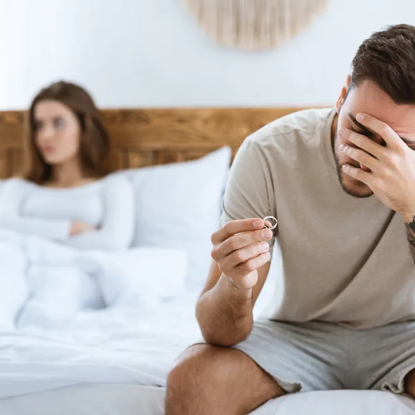 Distressed man holding a wedding ring on a bed with woman behind him