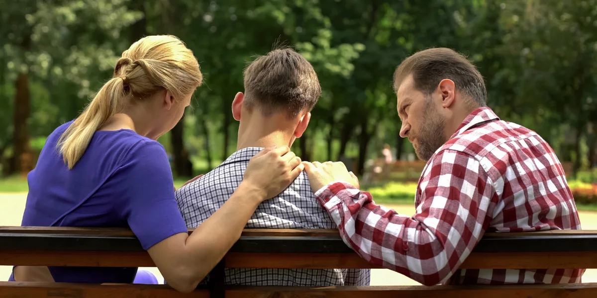 Parents on a park bench comforting a teenage boy seated between them